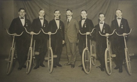 6 Männer in schwarzen Anzügen mit Fahrrädern, zwischen ihnen steht ein Mann ohne Farrad in einem braunen Anzug.