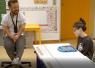 Ein kleinwüchsiger Mann und ein Kind mit Sprachcomputer in einem Klassenraum.
