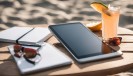 Foto: iPad, Notizbuch, Cocktail und Sonnenbrille auf einem Holztisch, dahinter Strand