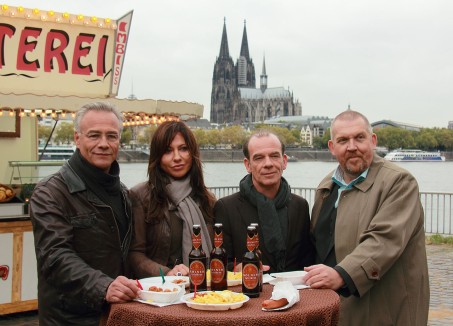 Vier Kommisare der Krimiserie Tatort stehen an einem Tisch neben einer Currywurstbude. Im Hintergund der Dom und der Rhein.