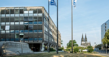 Ein viergeschossiges Bürogebäude, im Vordergrund eine große Wiese und ein mächtiger Baum. Im Hintergrund die Silhuette des Kölner Doms