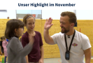 Ein Mann und ein Mädchen klatschen beieinander ein. Im Hintergrund steht ein weiteres Mädchen und eine Turnhalle ist zu sehen.