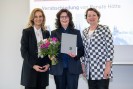 Drei Frauen in formeller Kleidung stehen lächeln nebeneinander. Die Frau in der Mitte hält einen Blumenstrauß mit pinken Blumen und eine graue Mappe in den Händen.
