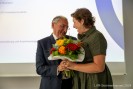 Man sieht eine lächelnde Frau in einem beigen Kleid, die einem lächelnden Mann im Anzug einen Blumenstrauß überreicht.