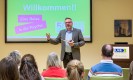 Man sieht einen Mann in blauem Hemd und grauem Anzug, der zu einem Publikum spricht. Im Hintergrund steht ein Bildschirm: Darauf steht "Willkommen Eine Reise durch die Psyche" auf grünem Grund.