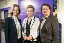 Drei Frauen in formeller Kleidung stehen vor einer violetten Ausstellungstafel der Gedenkstätte Brauweiler.
