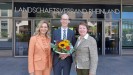 Ein Mann in dunkelgrauem Anzug und zwei Frauen stehen lächelnd nebeneinander. Der Mann hält einen bunten Blumenstrauß in den Händen.