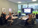 Eine Gruppe Menschen sitzt in einem Besprechungsraum an einem Tisch . Auf dem Tisch liegen Unterlagen. Im Hintergrund steht ein großer Bildschirm.
