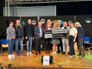 Man sieht eine Gruppe von Menschen. Im Hintergrund steht eine schwarze Stellwand mit bunten Peace-Zeichen drauf. Einige Personen der Gruppe halten ein Schild mit der Aufschrift: "Mitglied im Netzwerk Schule ohne Rassismus. Schule mit Courage."