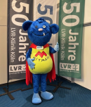 Der Mitmän, einer blauer Kobold in Superhelden-Montur, steht vor drei Fahnen mit der Aufschrift "50 Jahre LVR-Klinik Köln" und streckt den Daumen hoch.
