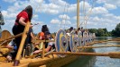 Ein großes hölzernes Boot mit gelb und blau bemalten Schilden an der Seite schwimmt auf dem Wasser. Eine Gruppe Menschen ist auf dem Boot zu sehen.