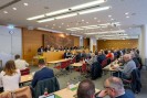 Eine Gruppe von Menschen sitzt in einem Konferenzraum, in dem viele Menschen an langen Tischen sitzen. An der Wand im Hintergrund sind viele Wappen zu sehen.
