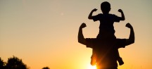 Silhouette eines Mannes mit Kind auf den Schultern im Gegenlicht der untergehenden Sonne