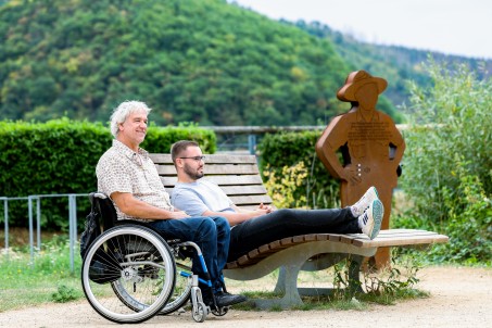 Zwei Männer genießen die Natur. Der eine Mann sitzt im Rollstuhl, der Mann neben ihm auf einer Parkbank.