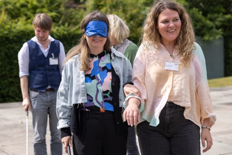 Eine Person mit Augenbinde und Stock, die von einer weiteren Person geführt wird.