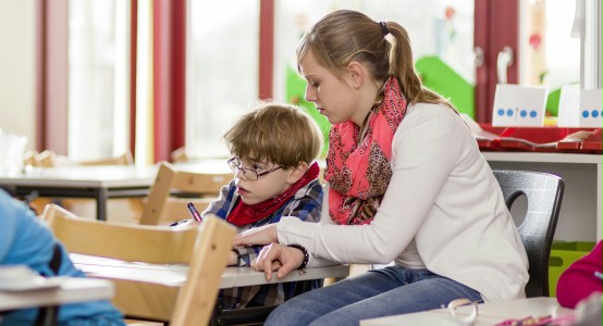 Das Foto zeigt einen kleinen Schuljungen während des Unterrichts. Er wird von einer jungen Frau beim Schreiben unterstützt. Foto: Dominik Schmitz, LVR-Zentrum für Medien und Bildung